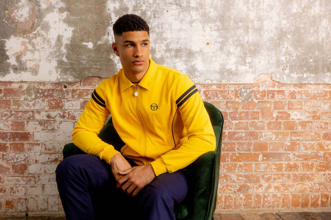 A model wearing The Sergio Tacchini Track Top in yellow, seated casually on a green chair against a textured wall.