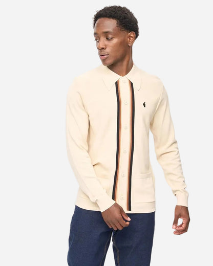 A man wearing a cream-colored knit button-up shirt with a vertical striped detail in black, brown, and orange, paired with dark blue jeans, photographed against a plain white studio background.