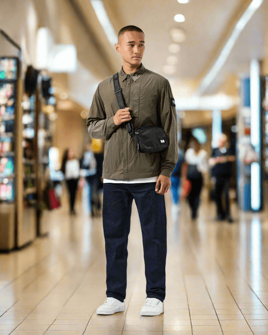 Man wearing Weekend Offender PORTER Overshirt in Castle Green with black shoulder bag and white sneakers in a shopping mall.